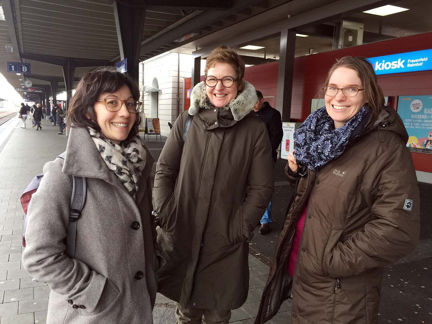 Drei Frauen lächeln und stehen am Bahnhof, umgeben von Reisenden und einem Kiosk im Hintergrund.
