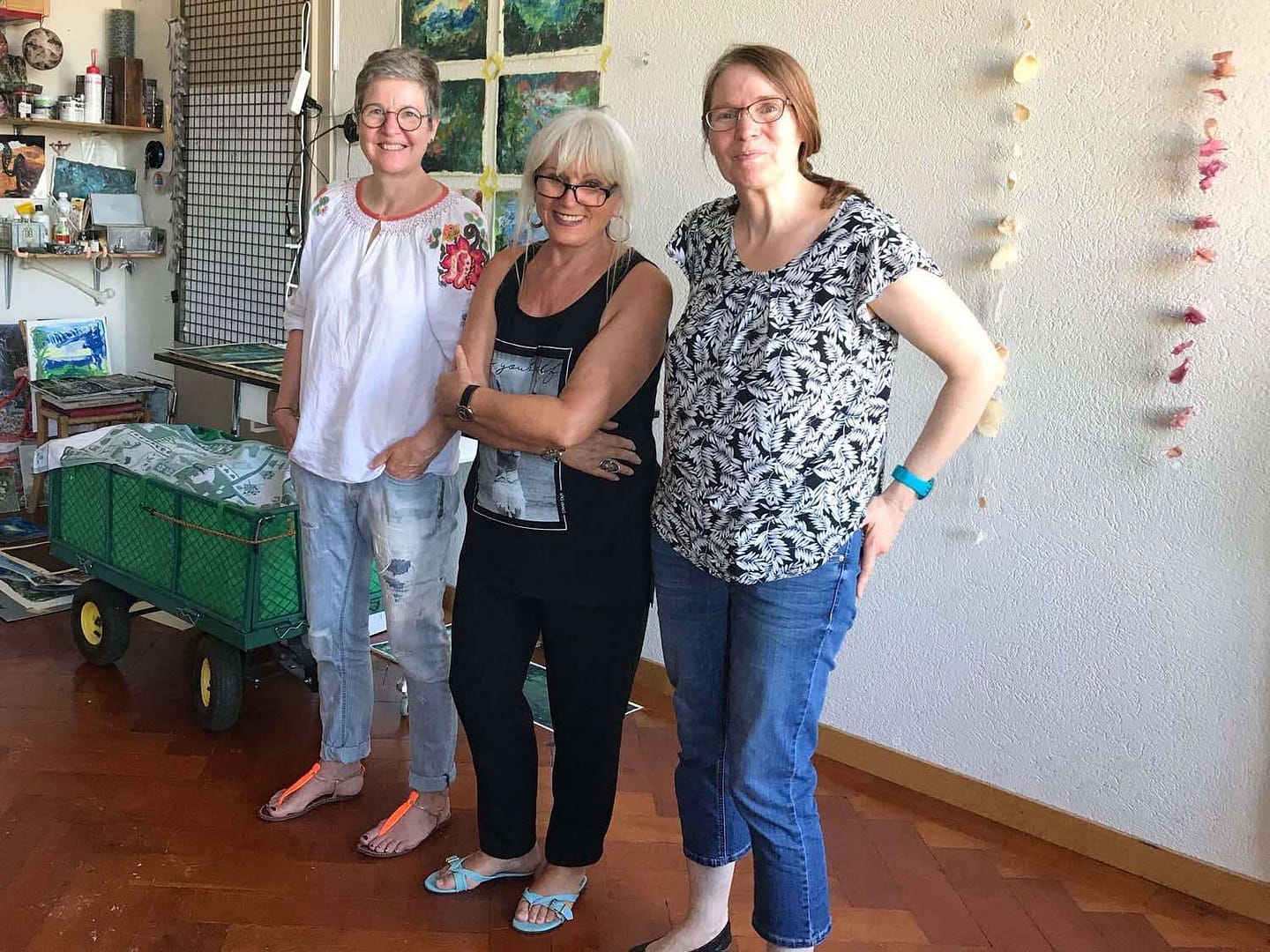 Drei Frauen stehen in einem kreativen Raum mit Kunstwerken im Hintergrund. Sie lächeln und posieren fröhlich für das Foto.