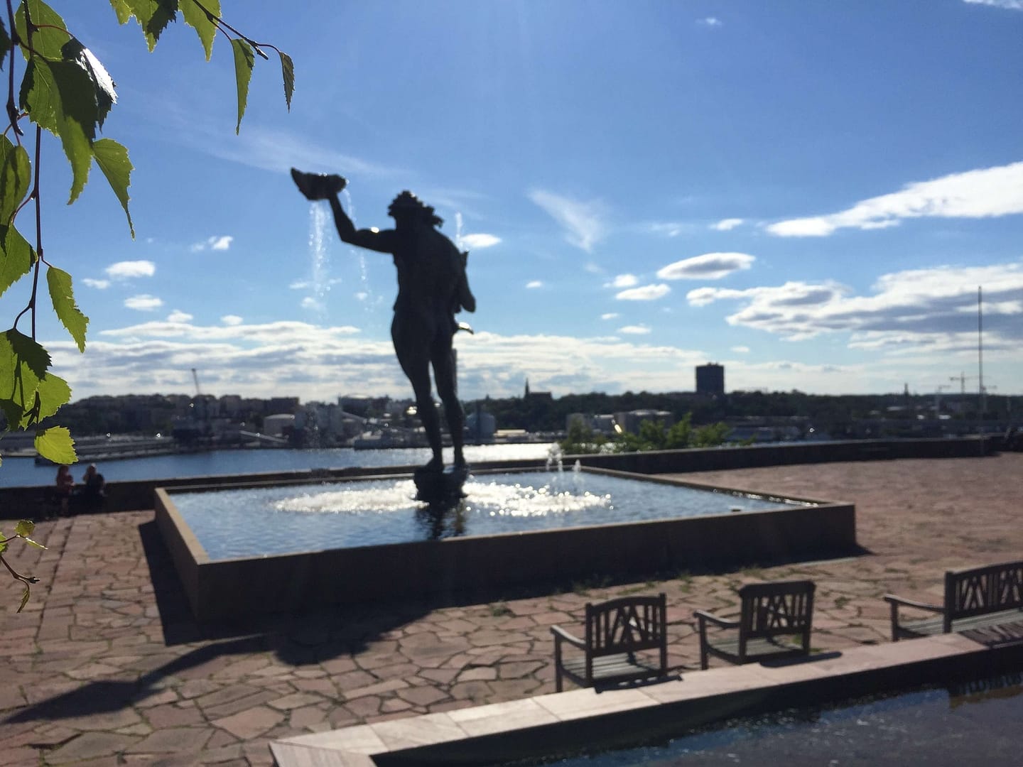Statue eines Mannes, der Wasser aus einem Gefäß gießt, vor einem Wasserbecken mit Blick auf die Stadt und den Himmel.