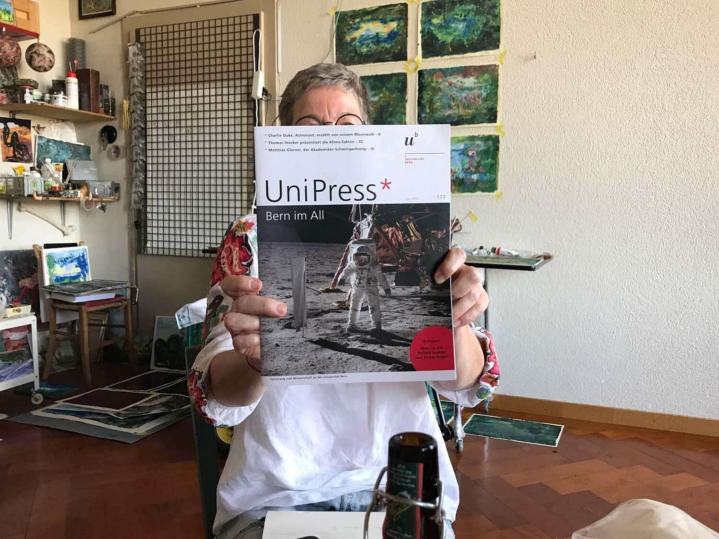 Eine Person hält die Ausgabe des Magazins 'UniPress' mit dem Titel 'Bern im All' in einem kreativen Raum mit Malereien im Hintergrund.
