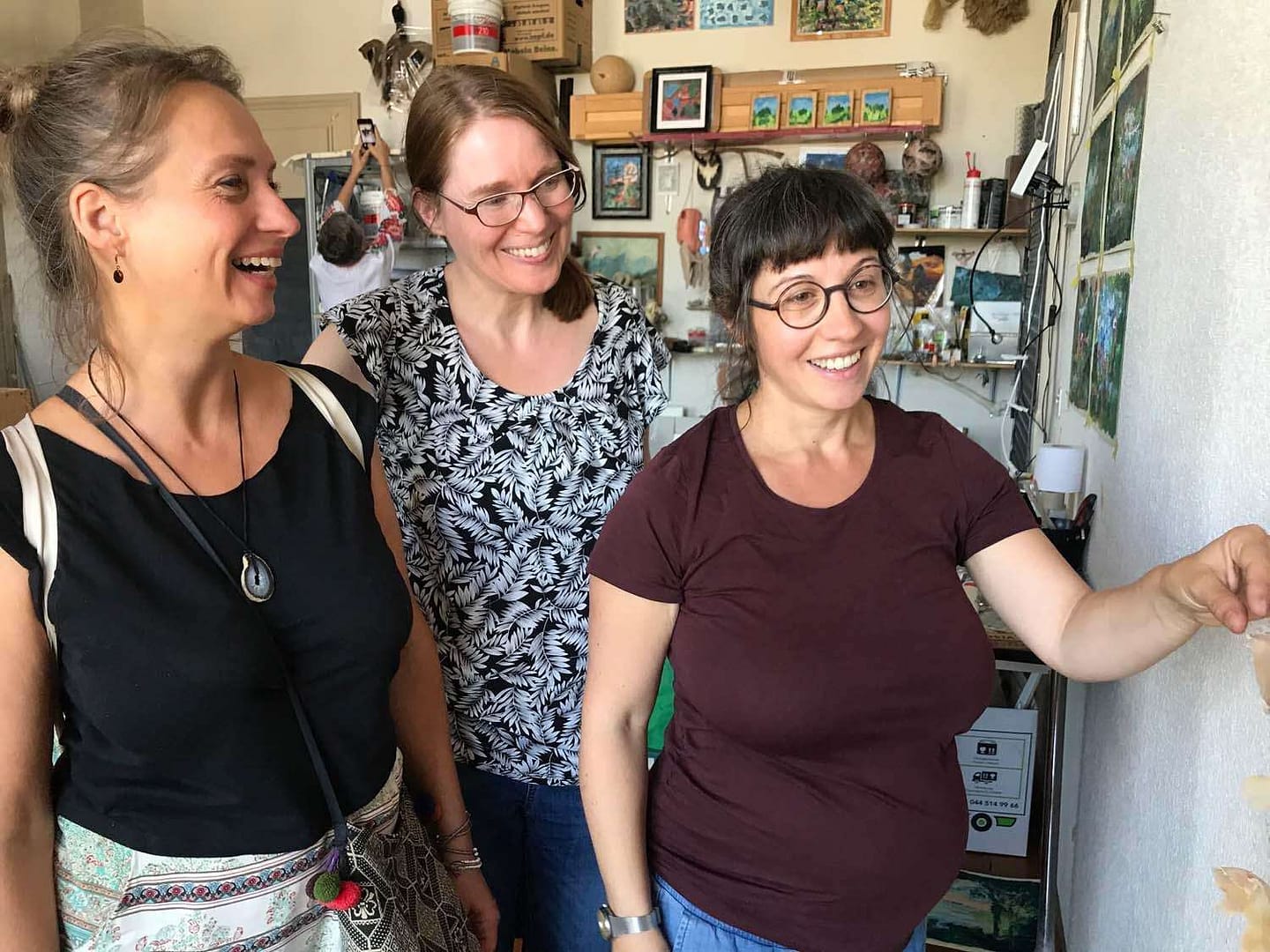 Drei Frauen lächeln und interagieren in einem kreativen Atelier, umgeben von Kunstwerken.
