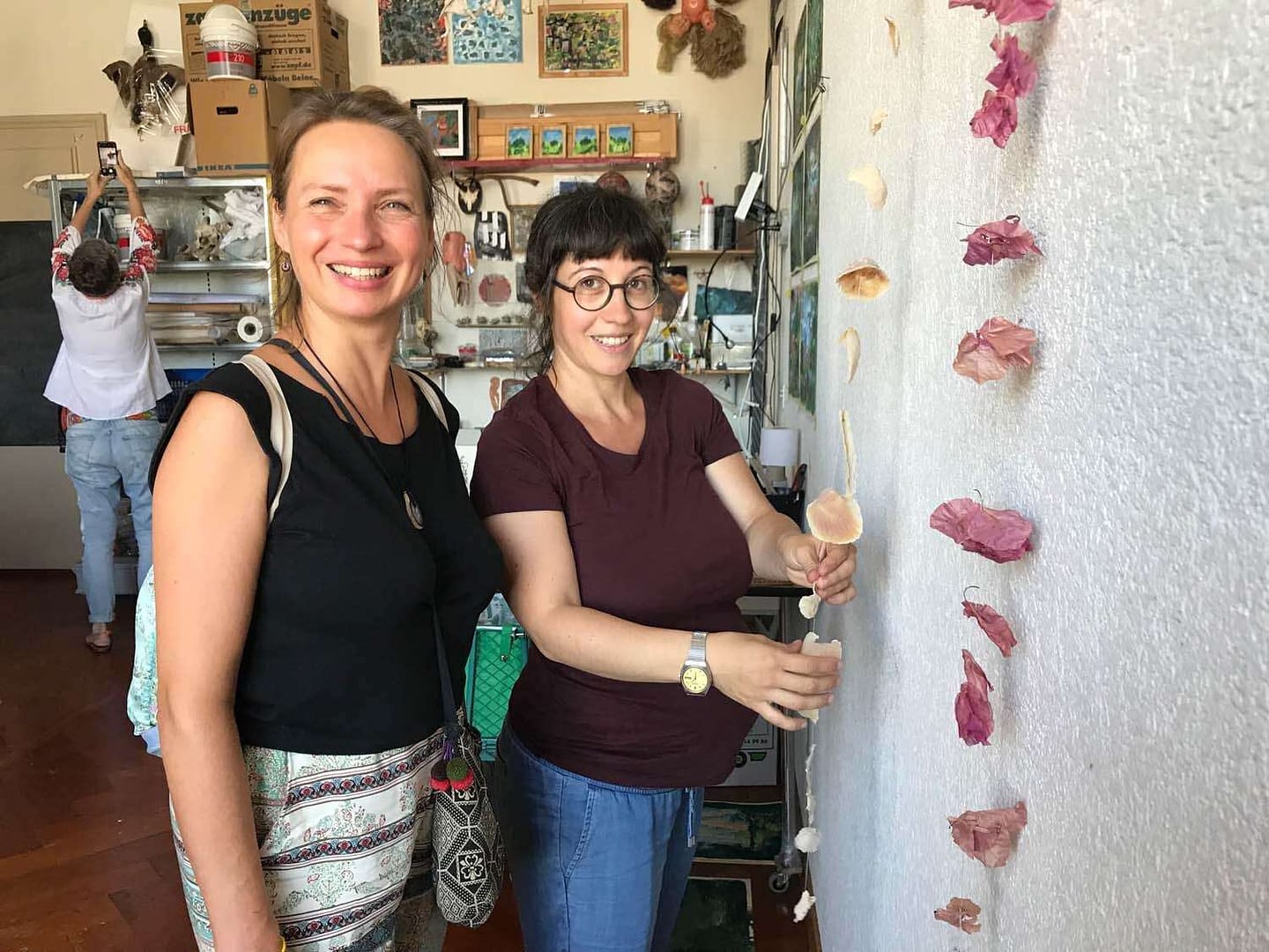 Zwei Frauen in einem kreativen Raum, eine mit einem Lächeln neben einer Wand, die mit kunstvollen Objekten dekoriert ist. Die andere Frau arbeitet an einem Handwerkstück. Im Hintergrund ist eine weitere Person zu sehen.