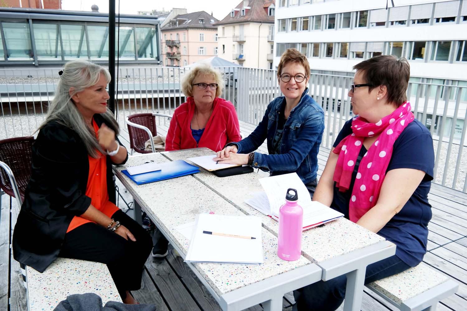 Vier Frauen sitzen an einem Tisch im Freien und diskutieren, umgeben von urbaner Architektur. Sie haben Bücher und Notizen vor sich.