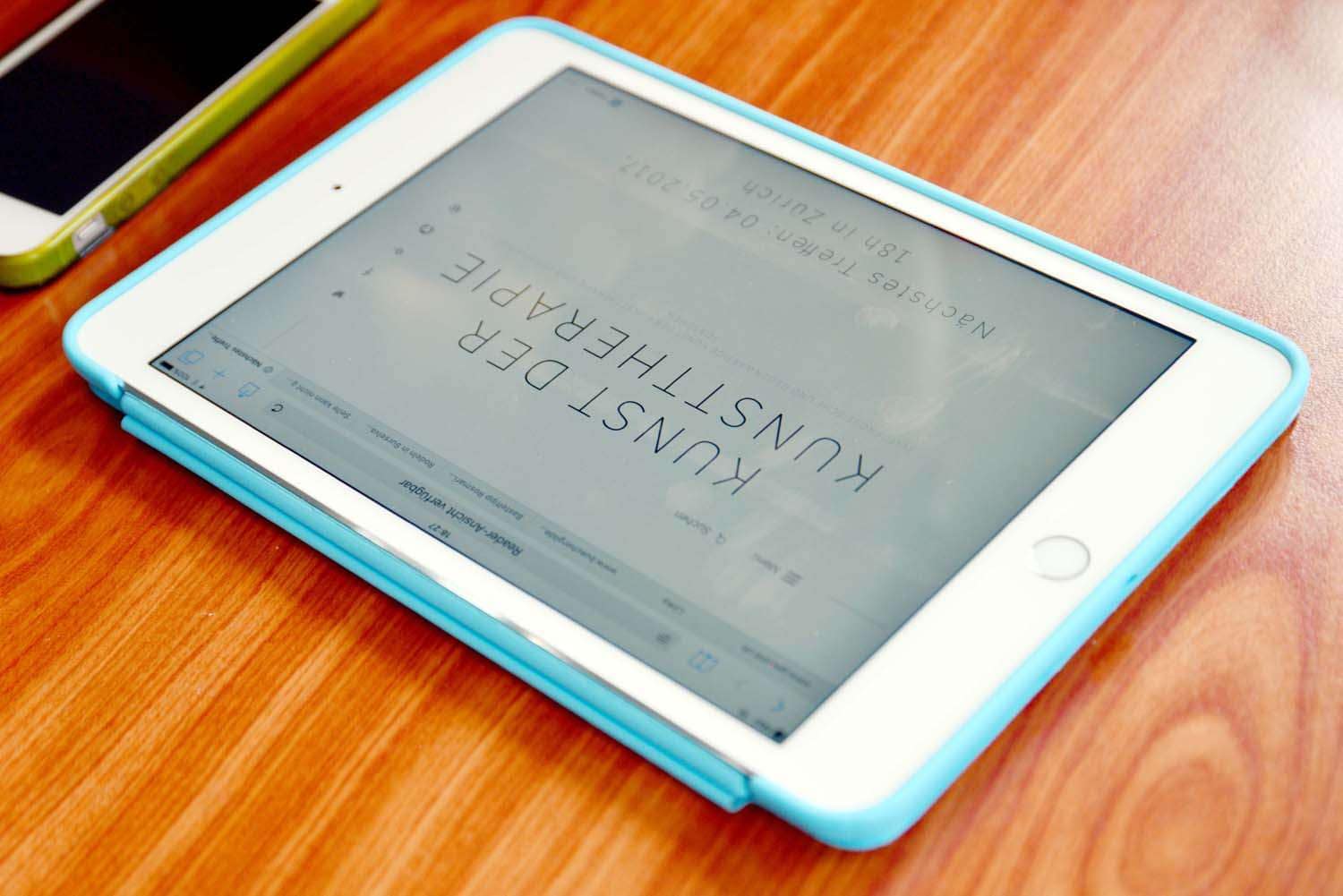 Ein Tablet mit einer blauen Hülle, das auf einem Holzschreibtisch liegt. Auf dem Bildschirm steht "KUNST DER KUNSTSTELLUNG."