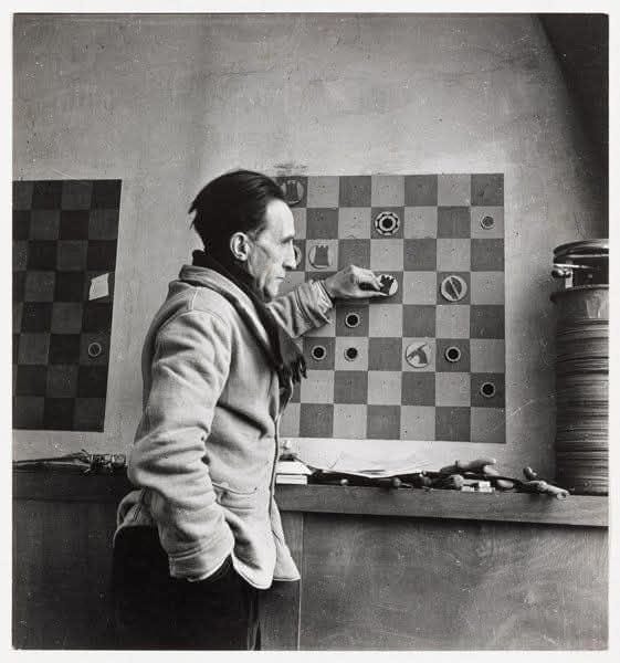 Ein Mann zieht eine Schachfigur auf einem Schachbrett, das an der Wand hängt. Im Hintergrund sind Werkzeuge und Materialien auf einem Tisch zu sehen.