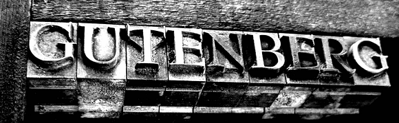 Schwarz-weißes Bild von einem Textblock mit dem Wort 'GUTENBERG' in großen, prägnanten Buchstaben, das auf einer alten Druckerpresse zu sehen ist.