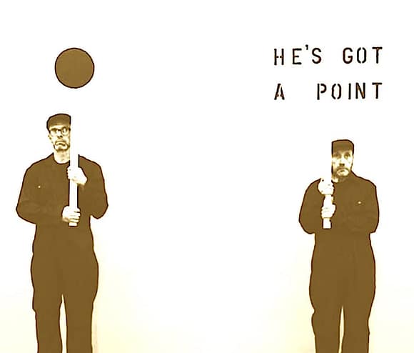 A performance artist holding a long stick with a circular object above their head, standing next to a sign that reads 'HE'S GOT A POINT', in a minimalistic setting.