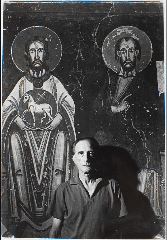 Ein Mann steht vor einem Wandgemälde mit zwei Heiligen, beide mit Heiligenscheinen, und hält ein Objekt in der Hand.