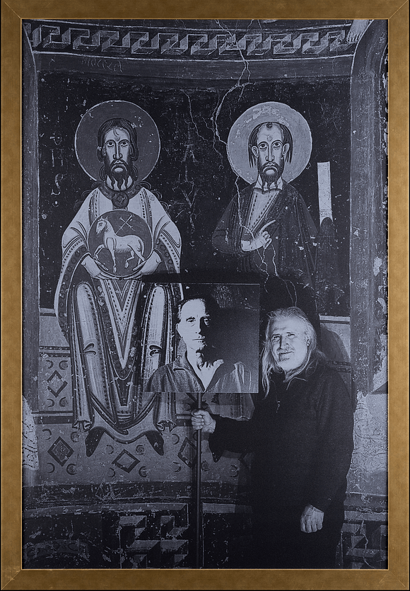 Eine Person hält ein Schild mit einem Porträt in einer minimalistisch gestalteten Umgebung, umgeben von zwei heiligen Darstellungen an der Wand.