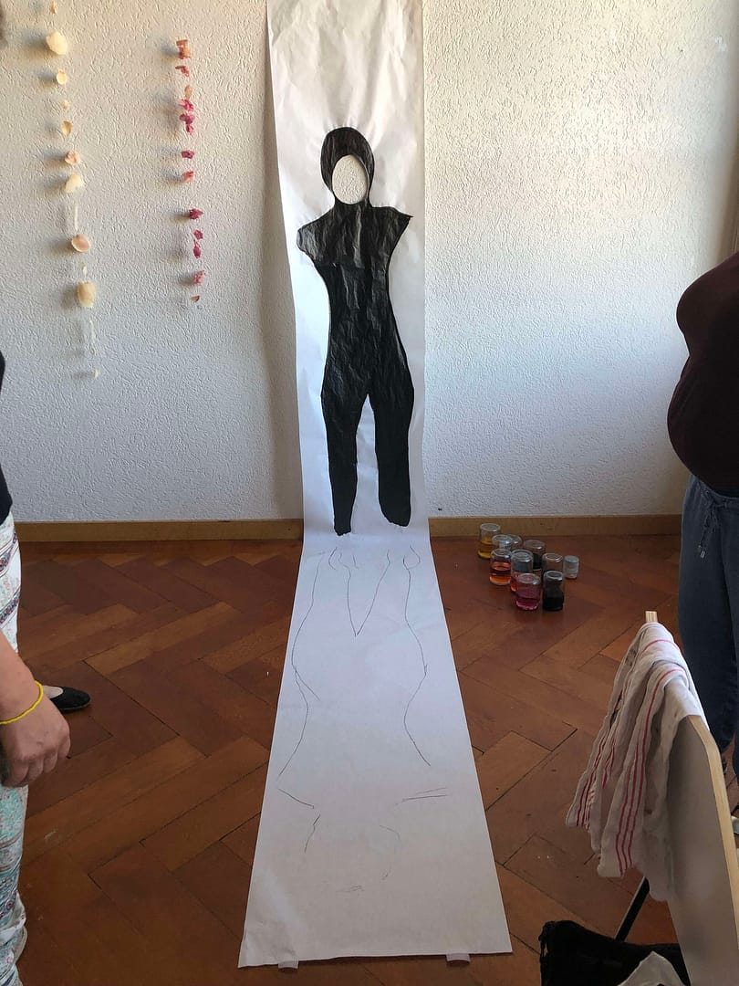 Eine kreative Installation auf einem weißen Papier, das eine menschliche Figur in Schwarz darstellt, die vor einer Wand steht. Unten sind umreißende Zeichnungen zu sehen, während farbige Töpfe im Hintergrund stehen.