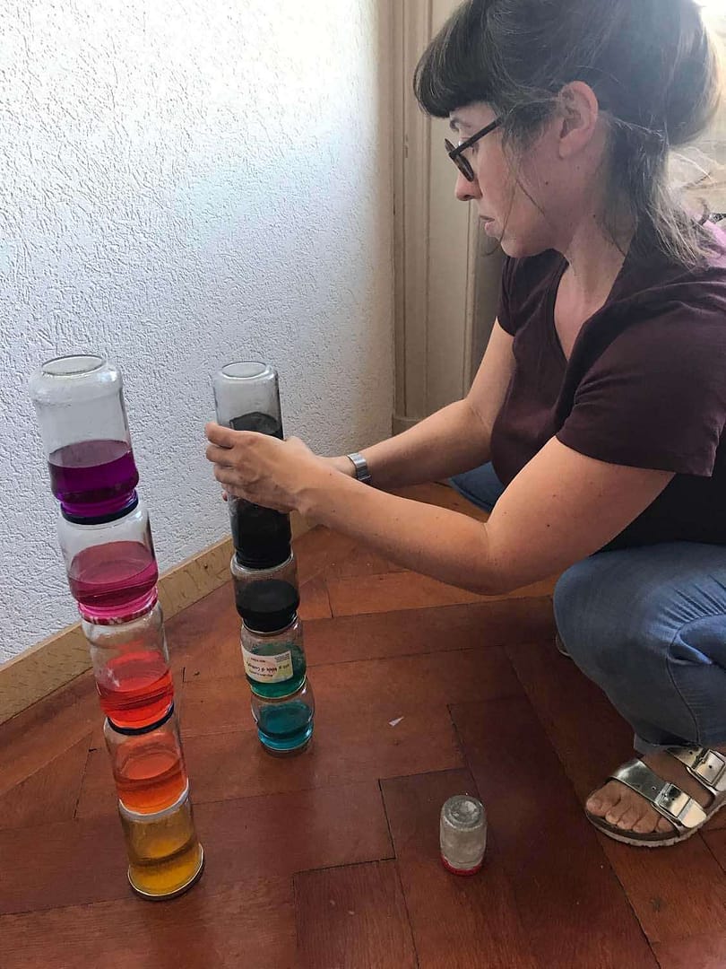 Eine Frau mit Brille, die bunte Gläser mit Flüssigkeiten stapelt, sitzt auf dem Boden.