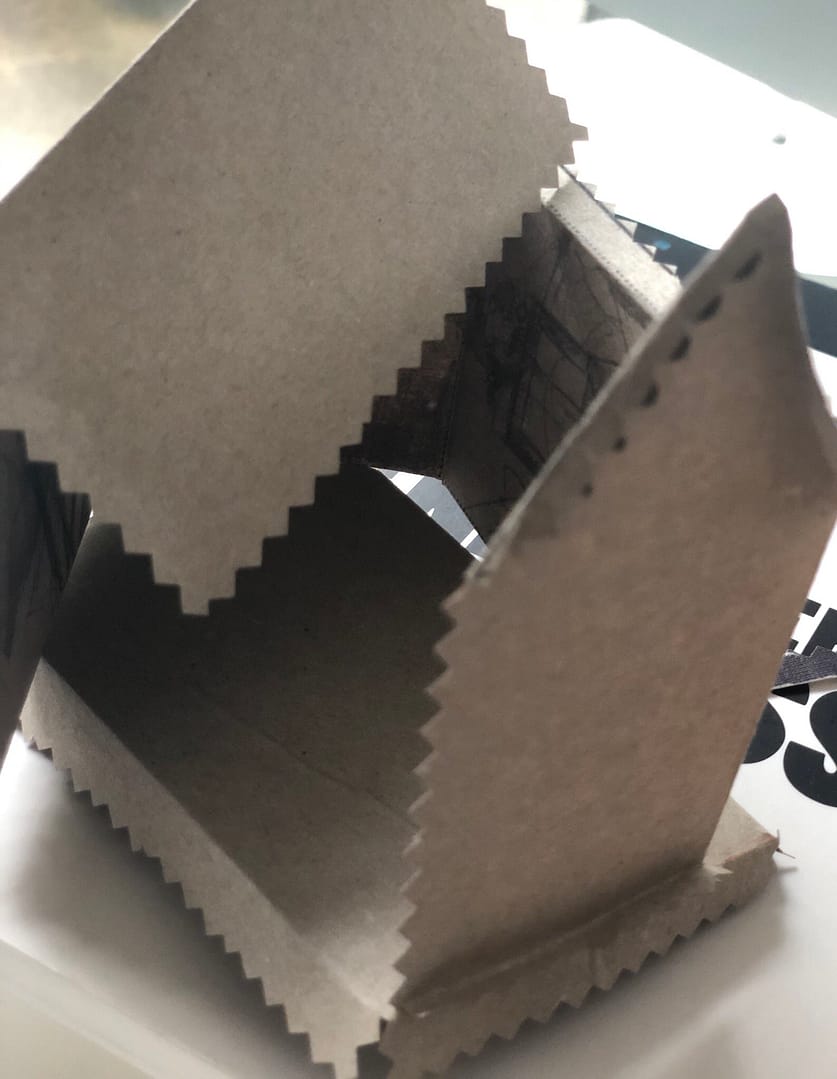 Eine geöffnetes, braunes Kartonmodell in Form eines Hauses, das auf einem Tisch liegt.