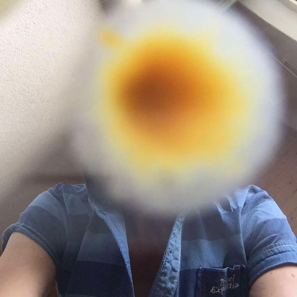 Ein unscharfes Foto, das eine Person zeigt, die mit einer Kamera oder einem Handy ein Selfie nimmt, während der Fokus auf einem gelben Objekt im Vordergrund liegt.